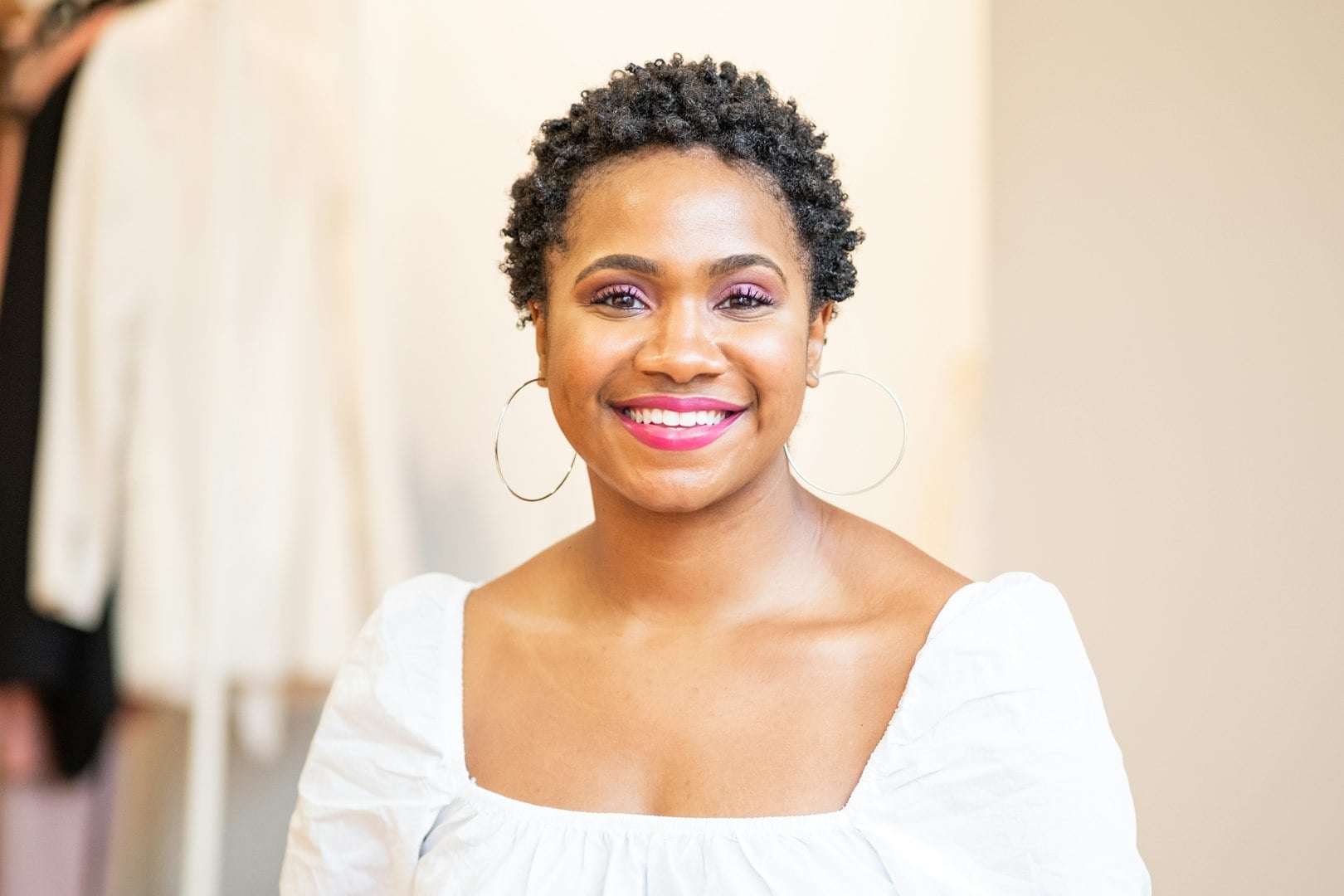 A woman with short brown hair smiles, wearing a light blue t-shirt, against a soft-focus neutral background.